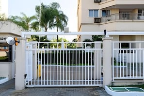 Una entrada protegida con un portón de metal blanco, una cámara de seguridad y un edificio con balcones al fondo.