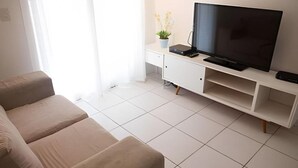 Ein Wohnzimmer mit einer Couch, einem Fernseher auf einem Schrank, einem kleinen Tisch mit einer Pflanze und einer weißen Kommode.