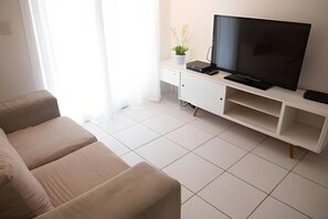 Un salon avec un canapé beige, un téléviseur à écran plat, une petite table sur laquelle est posée une plante, et un meuble blanc.