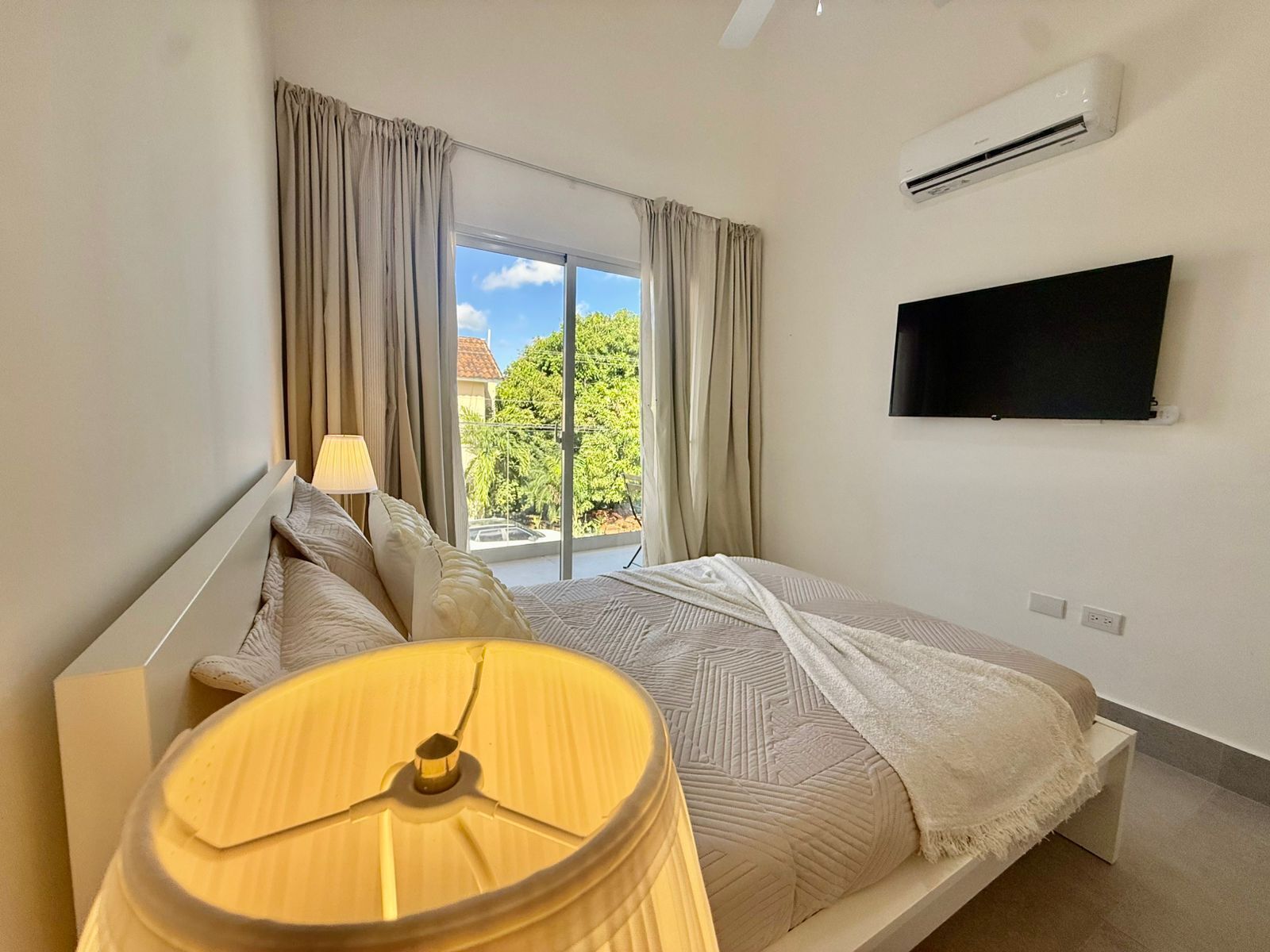 A bedroom with a bed, a TV, an air conditioner, and a window with curtains.
