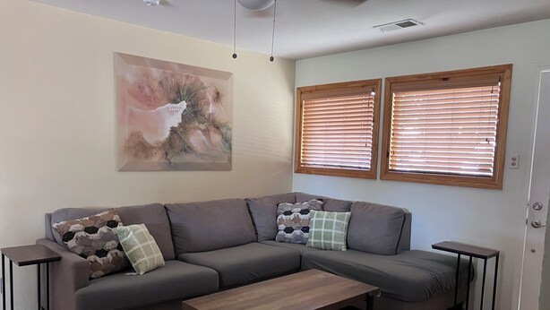 Een woonkamer met een grijze hoekbank, twee bijzettafels, een salontafel, een gekleurd tapijt en een schilderij aan de muur.