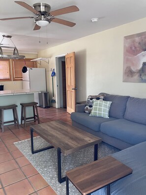 A living room with a sofa, a coffee table, a ceiling fan, a kitchen area with a refrigerator, and a door leading to another room. - North Valley Charmer (Albuquerque)