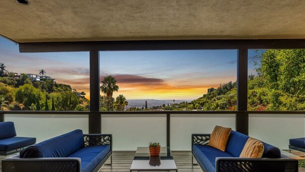 Ein Balkon mit zwei Sofas, einem kleinen Tisch und Blick auf einen Sonnenuntergang über einer hügeligen Landschaft.
