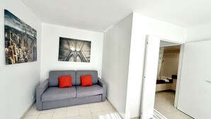A living room with a grey sofa, two red cushions, a cityscape painting, and a bridge painting on the wall. - Antibes - Studio for 2 people - Parking - Swimming pool - Air conditioning (Antibes)