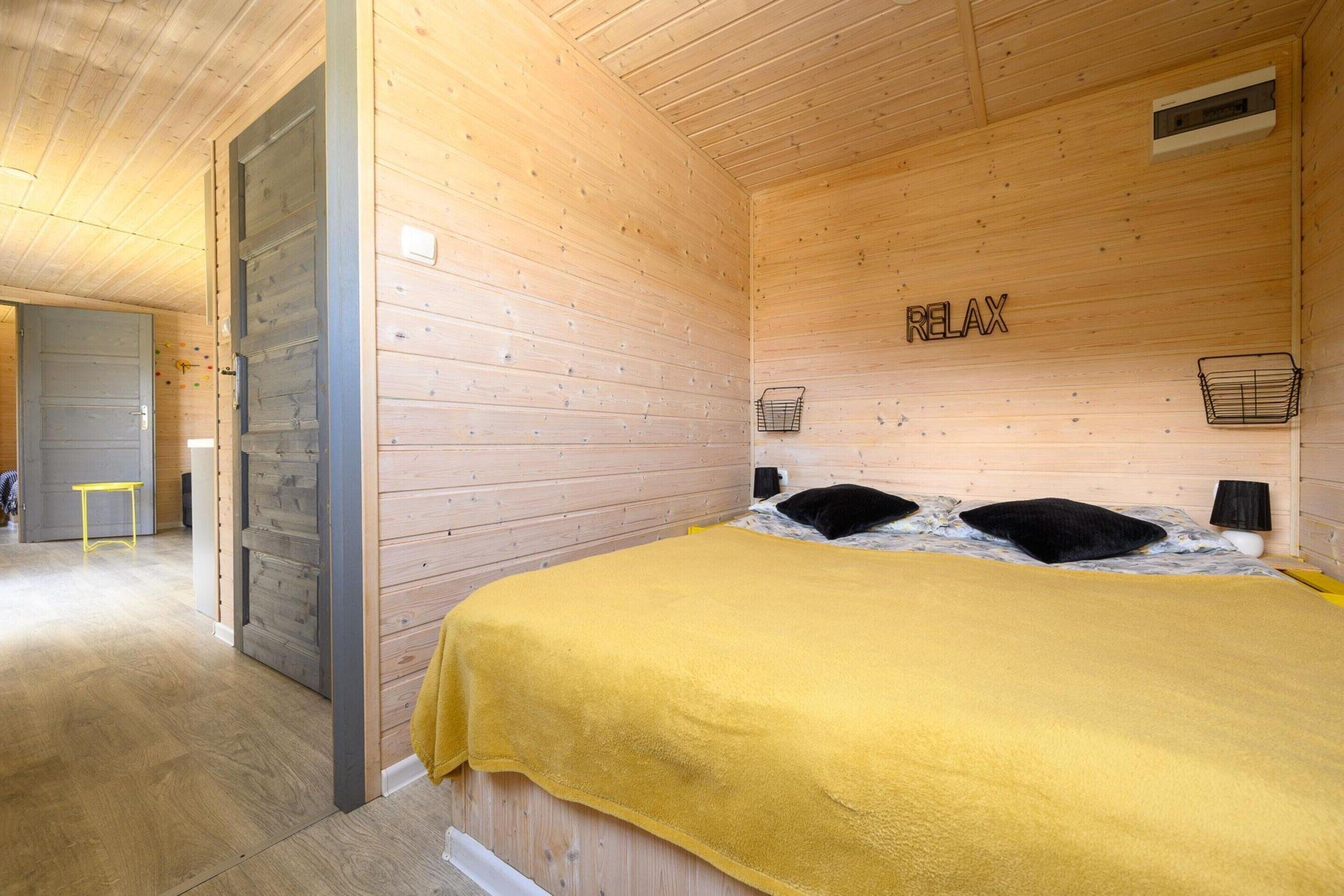 Une pièce aux murs en bois, un lit recouvert d’une couverture jaune, deux oreillers noirs, une petite table de chevet et une étagère fixée au mur.