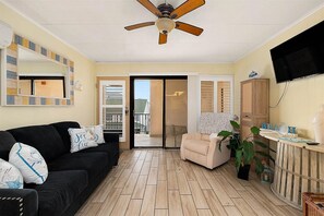Ein Wohnzimmer mit schwarzem Sofa, beigeem Sessel, Deckenventilator, Fernseher und Holzboden.