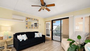 Quarto com sofá preto, ventilador de teto, espelho, janela com persianas e uma planta.