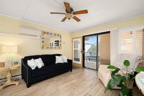 Ein Zimmer mit einer schwarzen Couch, einem Deckenventilator, einem Spiegel, einem Fenster mit Jalousien und einer Pflanze.