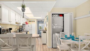 Cozinha compacta com armários brancos, área de refeições com cadeiras brancas, planta pendente e máquina de lavar embutida.
