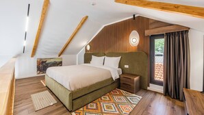 Una camera da letto con soffitto spiovente, travi in legno, un letto con testiera, comodini, un tavolino da notte, un tappeto decorato e una finestra con tende.
