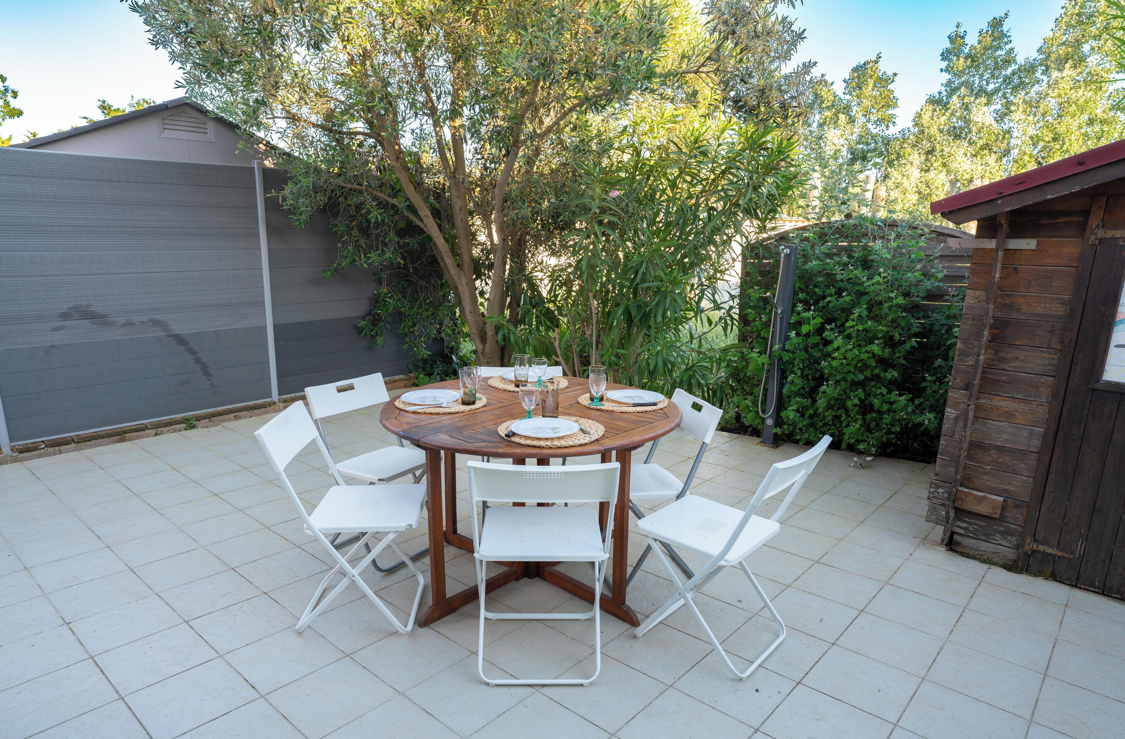 Una mesa redonda de madera con sillas blancas, rodeada de follaje y un cobertizo en un patio trasero.