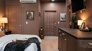 A bedroom with a bed, bedside table, lamp, air conditioning unit, and a TV mounted on the wall. - Moody Maison Studio | Near Downtown & Beaches (Traverse City)
