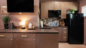 A kitchen with wooden cabinets, a black refrigerator, a microwave, and a flat-screen TV mounted on the wall. - Moody Maison Studio | Near Downtown & Beaches (Traverse City)