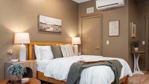 A bedroom with a wooden bed, two bedside lamps, a nightstand with a vase, a wall-mounted air conditioner, and a framed picture. - Sage Solace Studio | Near Downtown & Beaches (Traverse City)