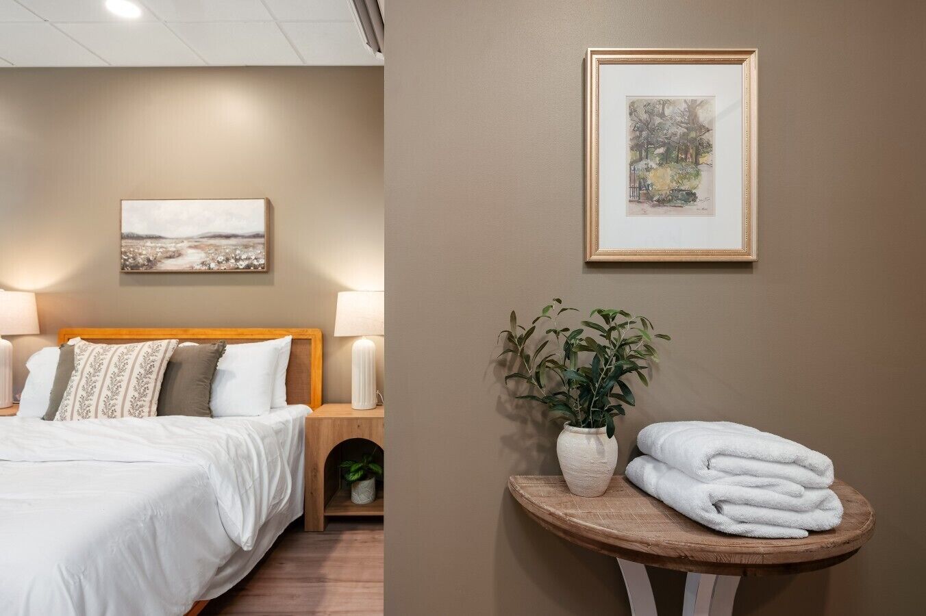A hotel room with a bed, bedside lamps, a wall-mounted painting, a potted plant, and a round wooden table with towels.