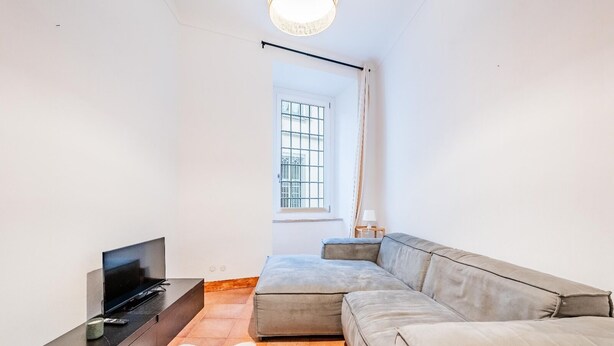 Una sala de estar moderna con una televisión de pantalla plana, un sofá gris, una mesita de noche con una lámpara y una ventana con rejas.