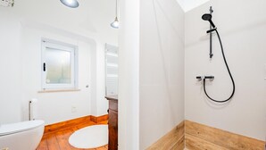 Un baño con inodoro, suelo de madera, alfombra blanca, ventana y ducha con base de madera.