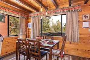 A wooden cabin interior with a dining table set for four, a fan, and a view of trees through large windows. - Peek-A-Boo Lake View, Dog Yard, Near Village (Big Bear Lake)