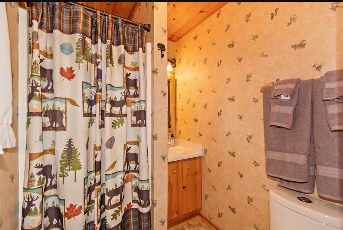 Et badeværelse med bruseforhæng med elge og skovmotiv, et træskab og et toilet med håndklæder hængende på væggen.