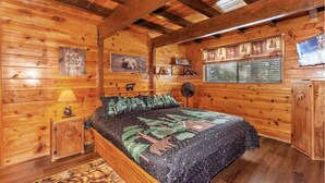 Ein Schlafzimmer in einer Holzhütte mit einem Bett, einem Nachttisch, einer Lampe, einem Ventilator, einem Fenster mit Vorhängen sowie einer Wand mit einem Bär-Bild und einem Schild.