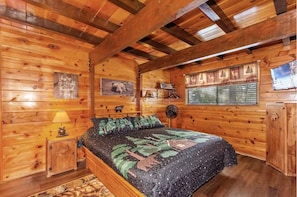 A wooden cabin bedroom with a bed, a nightstand, a lamp, a fan, a window with curtains, and a wall with a bear picture and a sign. - Peek-A-Boo Lake View, Dog Yard, Near Village (Big Bear Lake)