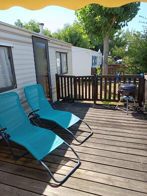 Eine Holzterrasse mit zwei grünen Liegestühlen, einem Grill und einem Holzzaun.