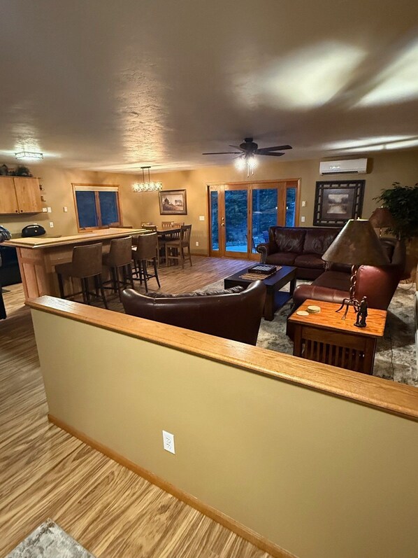 A spacious living area with a dining table, chairs, a sofa, and a coffee table. There is a kitchen area with cabinets and a countertop. A ceiling fan is visible, and the room has a warm color scheme. - Spacious Hillside Cabin Near Slopes (Lead)