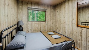 A single bed with a headboard, a bedside lamp, and a window with a view of trees. - 3-Bedroom Cottage #5 (Nobel)