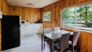 A wooden-paneled room with a glass table and chairs, a black refrigerator, and a window with a view of trees. - 3-Bedroom Cottage #5 (Nobel)