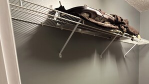 Une pièce avec un mur gris, un plancher en bois, une étagère en métal avec des vêtements et un sac posé sur le sol.