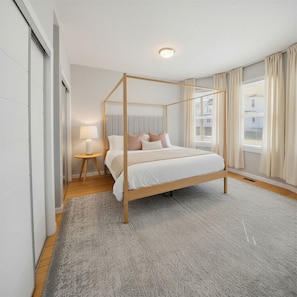 Ein modernes Schlafzimmer mit Himmelbett, Nachttisch mit Lampe, großem Fenster mit Vorhängen und Blick nach draußen.