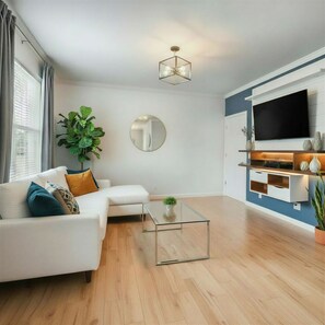 Ein modernes Wohnzimmer mit einem weißen Sofa, einem Glastisch, einem Flachbildfernseher und Holzboden.