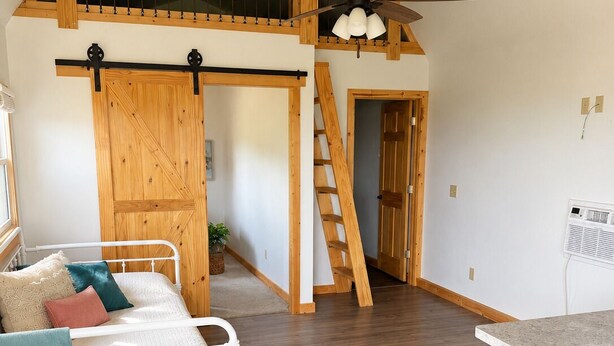 A room with a loft bed, a wooden ladder, a ceiling fan, and a kitchen area with a countertop. - Spacious Cottage 4 with Loft, Full Kitchen & Stunning Views (Lake Andes)