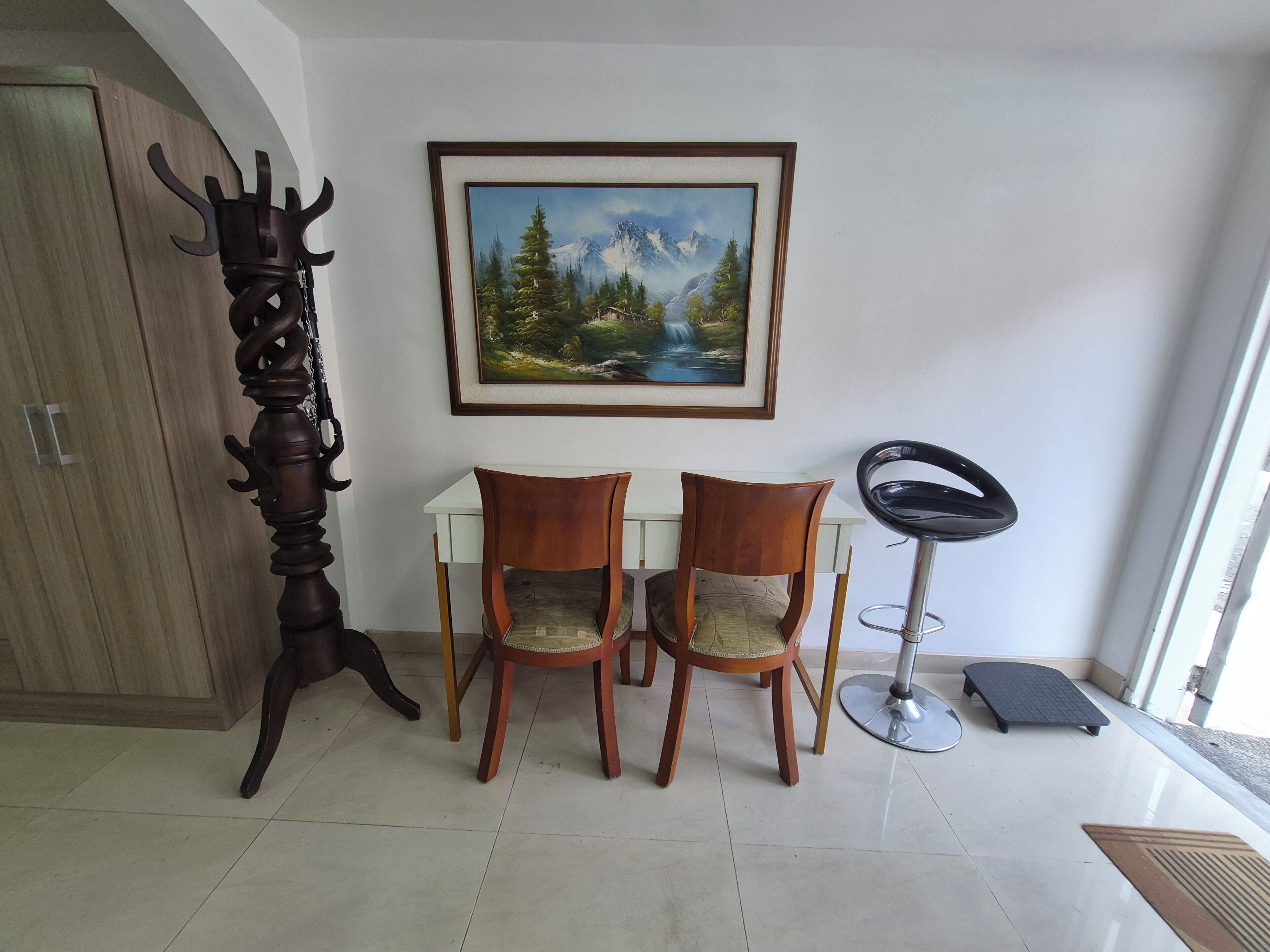 A room with a wooden cabinet, two wooden chairs, a framed painting of a mountain landscape, and a black chair with a metal base.