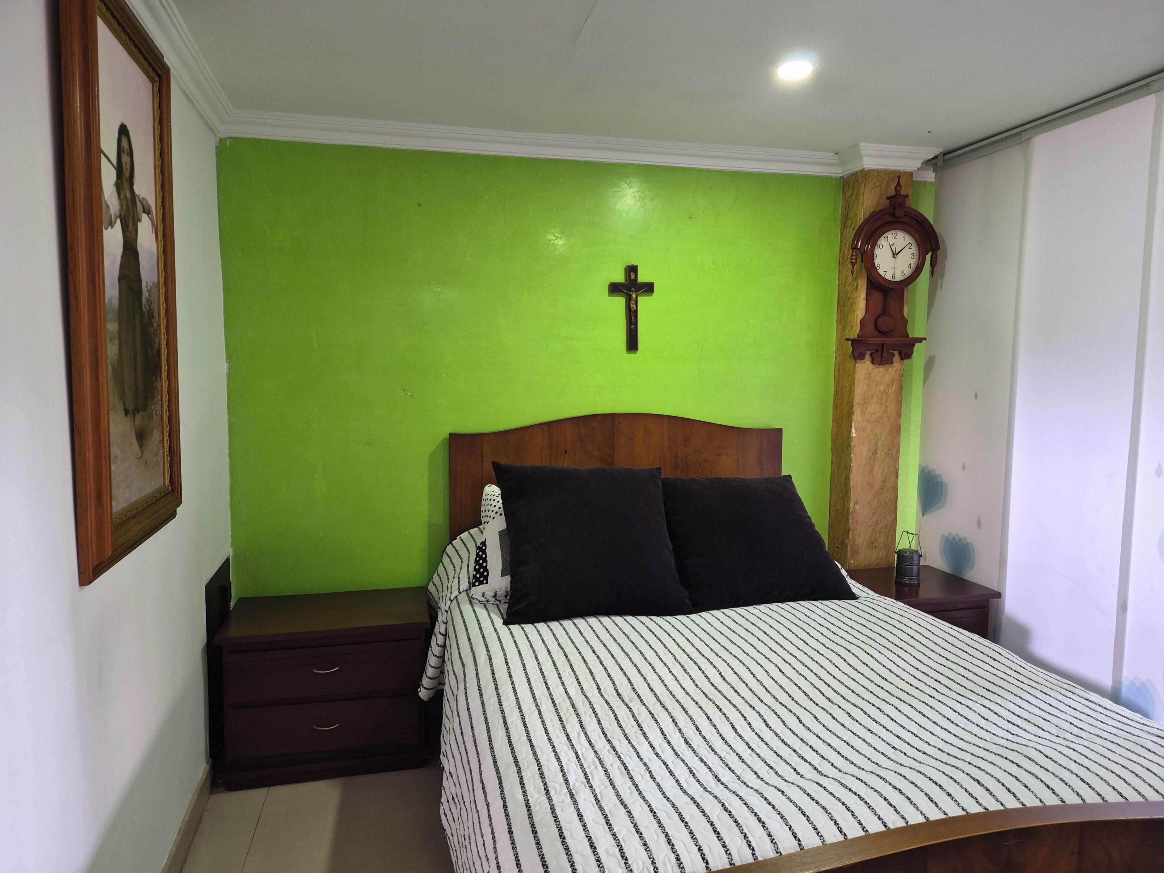 A bedroom with a green wall, a wooden headboard, a bed with striped bedding, a nightstand, a small table with a clock, and a framed picture on the wall.