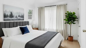 A bedroom with a bed, a nightstand, a lamp, a window with curtains, a plant, and a painting on the wall.