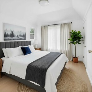 A modern bedroom with a bed, a bedside table, a lamp, a window with curtains, a plant, and a painting on the wall.