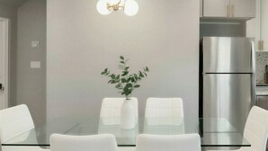 A modern dining area with a glass table, white chairs, a potted plant, and a stainless steel refrigerator.