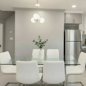 A modern dining area with a glass table, white chairs, a potted plant, and a stainless steel refrigerator.