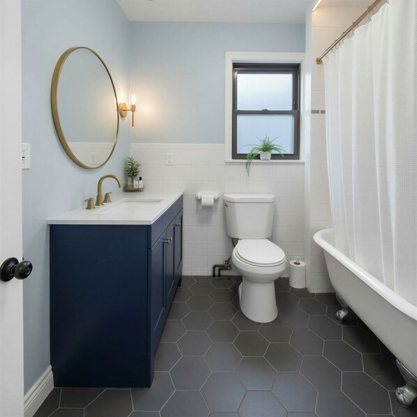 Une salle de bain avec un meuble-lavabo bleu foncé, des toilettes blanches, une fenêtre avec une plante et une baignoire blanche avec un rideau de douche.