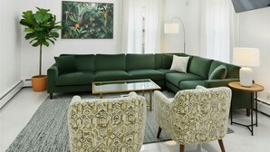 A modern living room with a green sofa, patterned armchairs, a coffee table, a large plant, and a tropical-themed painting.
