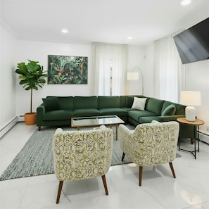 A modern living room with a sectional sofa, two patterned armchairs, a coffee table, a large potted plant, a wall art piece, and a floor lamp.