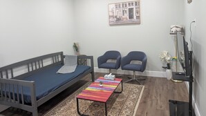 Quarto pequeno e bem iluminado com uma cama de solteiro, duas cadeiras, uma mesa colorida, uma TV montada e um quadro na parede.