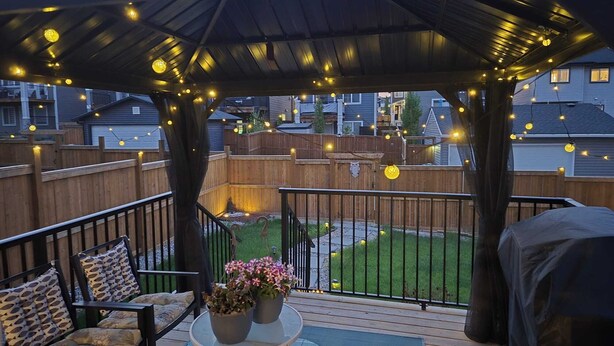 Um terraço coberto ao ar livre com luzes de cordão, uma mesa com plantas em vasos e uma área de estar com almofadas.