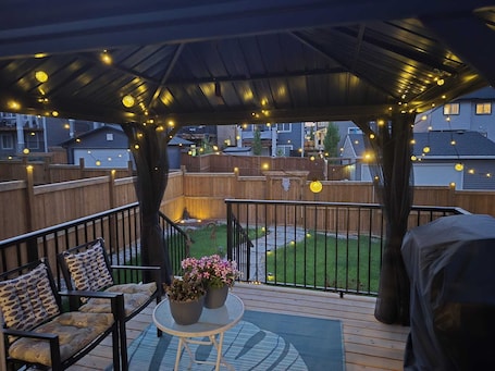 A covered outdoor deck with string lights, a table with potted plants, and a view of a backyard.. Spacious and cozy 1 Bedroom suite close to Calgary,Canmore and Banff Ntnl park.