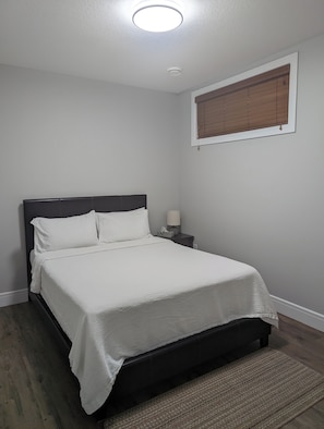 Un dormitorio con cama, mesita de noche, lámpara, ventana con persianas y luz de techo.