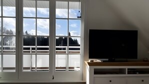 Una habitación con un ventanal grande, un televisor de pantalla plana sobre un soporte de madera y un armario blanco.