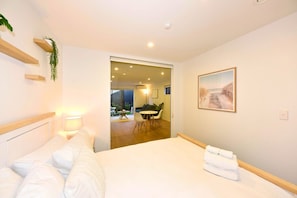 A hotel room with a bed, bedside table, lamp, and a painting. There is a door leading to a balcony with a table and chairs. - Contemporary City Stay with Wellness Access (Christchurch)