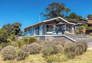 A modern house with a glass facade, a wooden deck, and a staircase leading to the entrance. - The Belle House – Where Sea Meets Sky (Waiheke Island)
