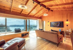 A spacious living room with a large window, a sofa, a television, and a wooden ceiling. - The Belle House – Where Sea Meets Sky (Waiheke Island)
